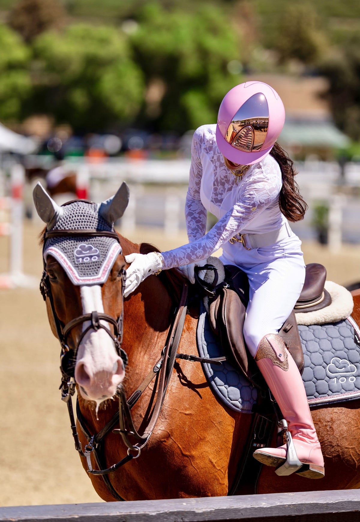 A woman in white equestrian riding attire with a pink helmet sits on a brown horse in an outdoor riding arena.