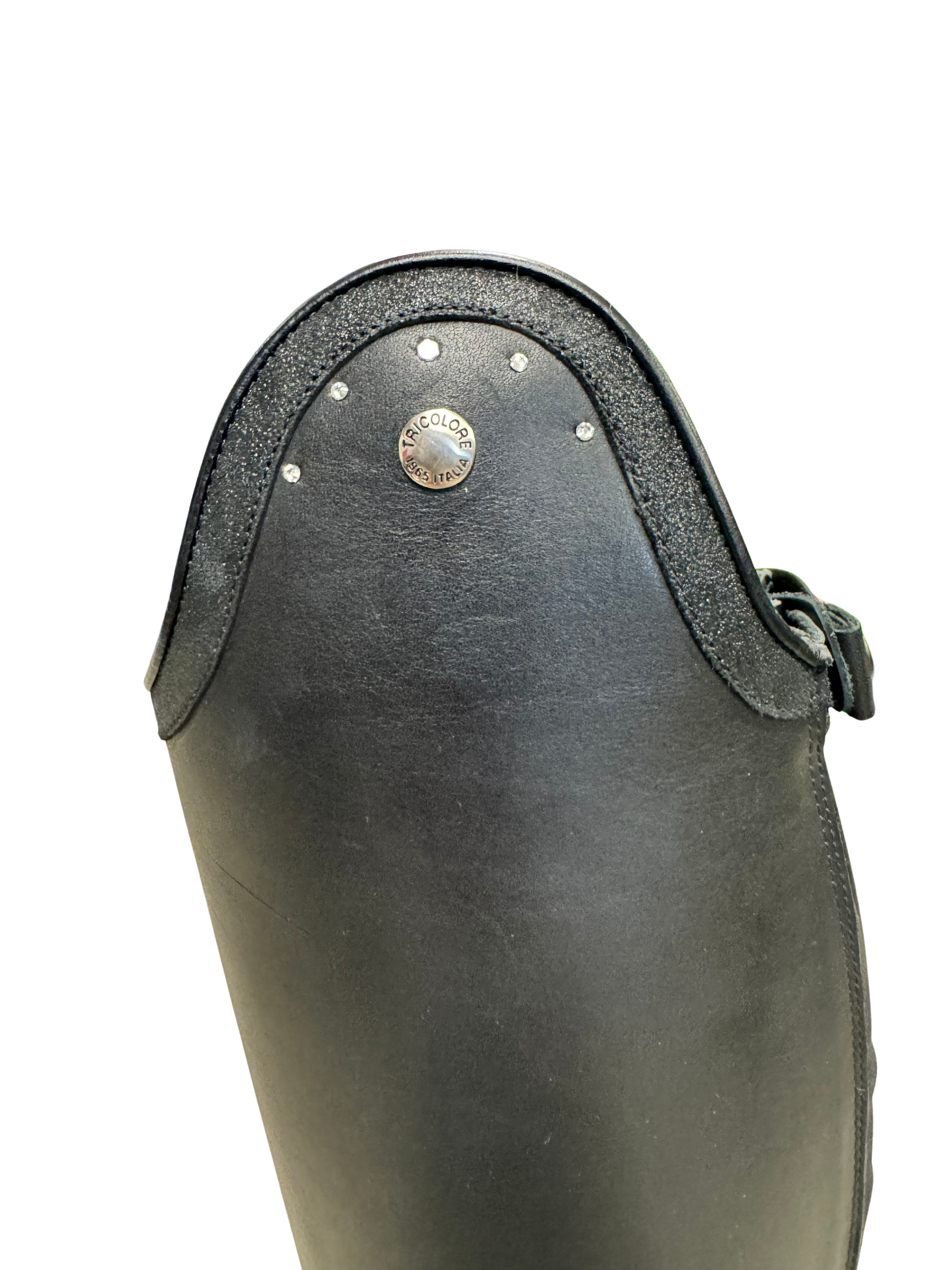 Close-up of a black leather riding boot showing the top front panel with a silver-toned circular medallion and small metal studs, on a white background.