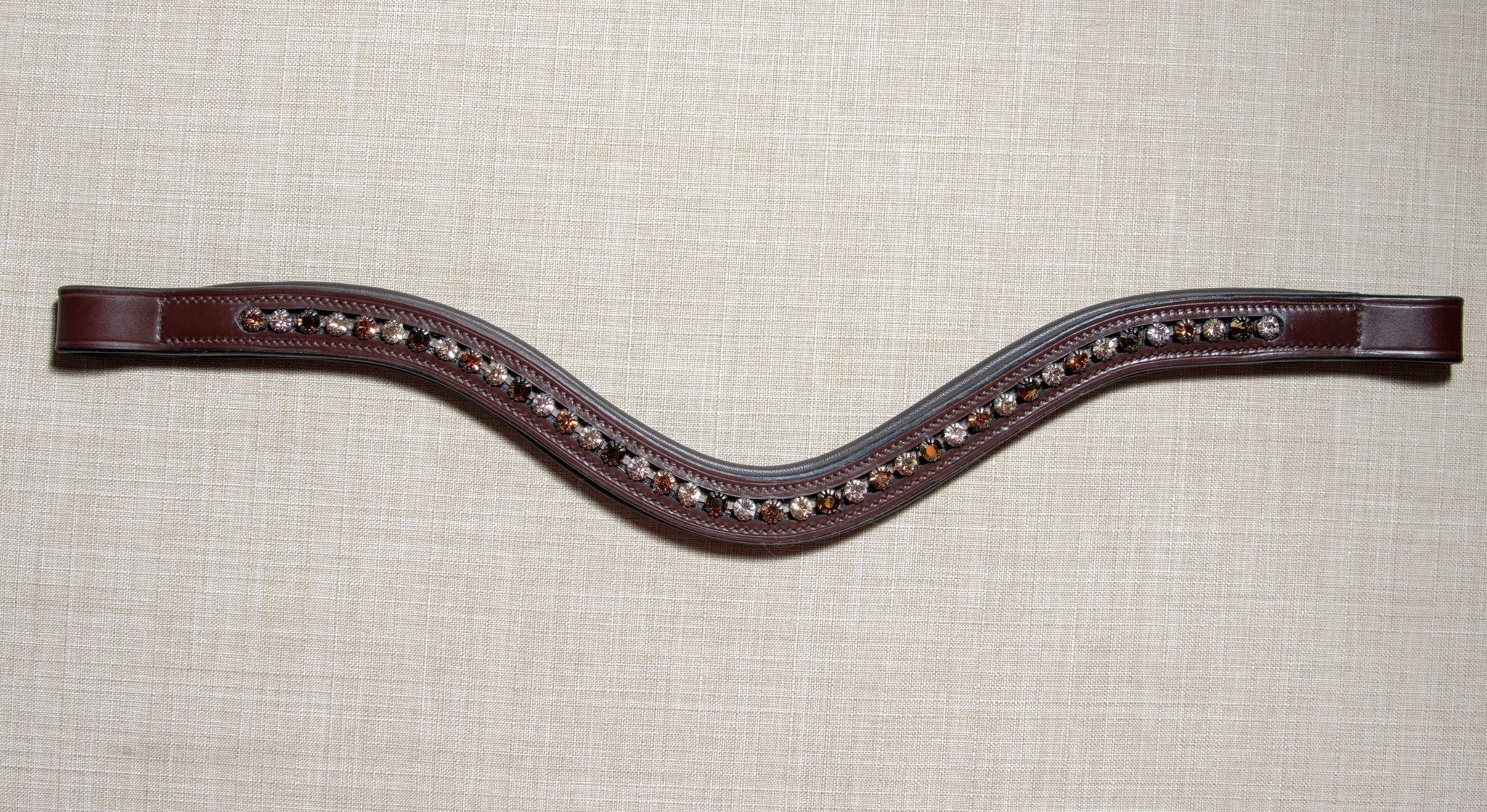 A dark brown leather horse bridle brow band with a wavy center and rows of metallic studs, displayed flat on a textured beige fabric background.