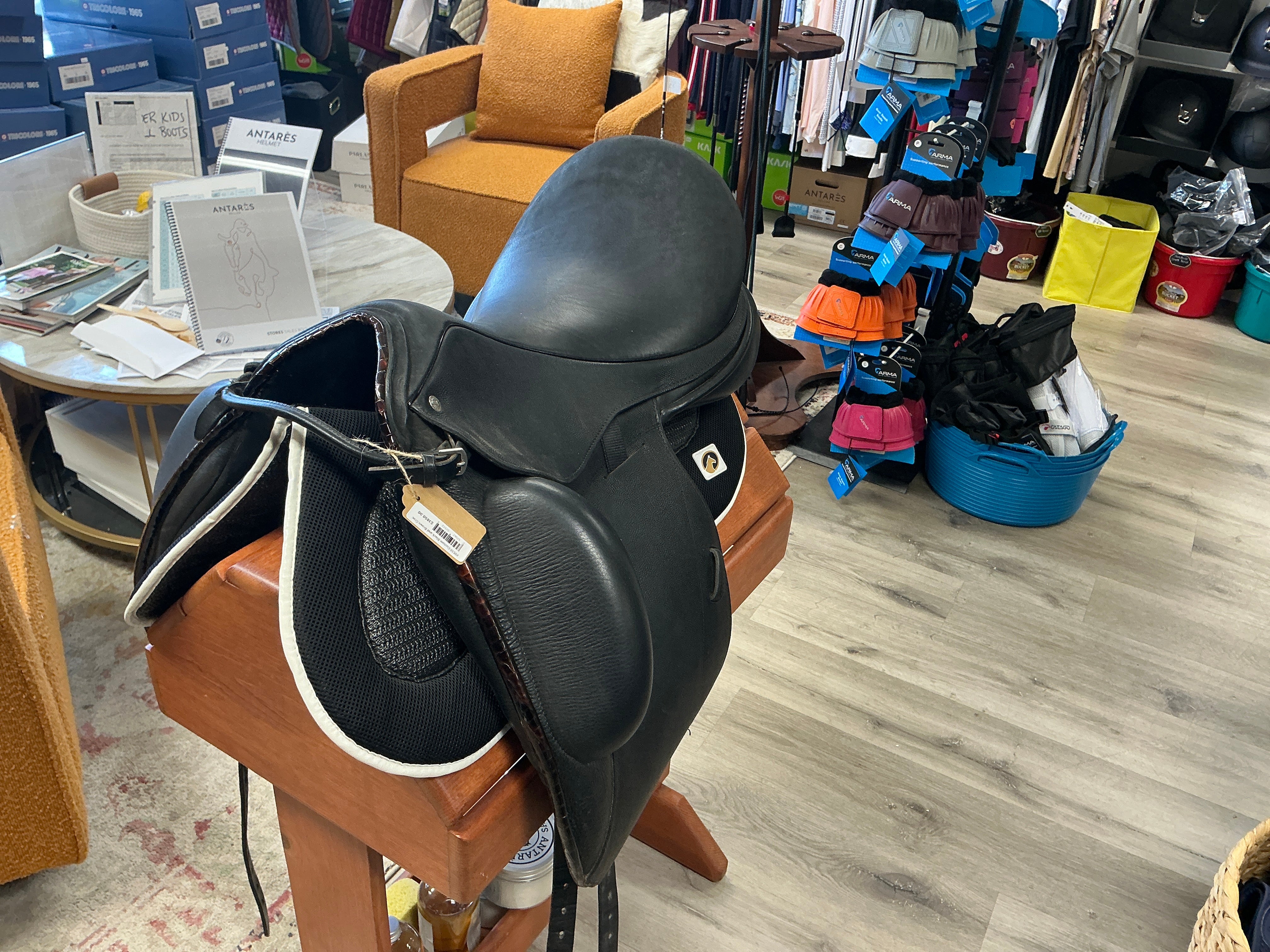 Black leather Custom Saddelry saddle on a wooden stand in a store setting with various items in the background.